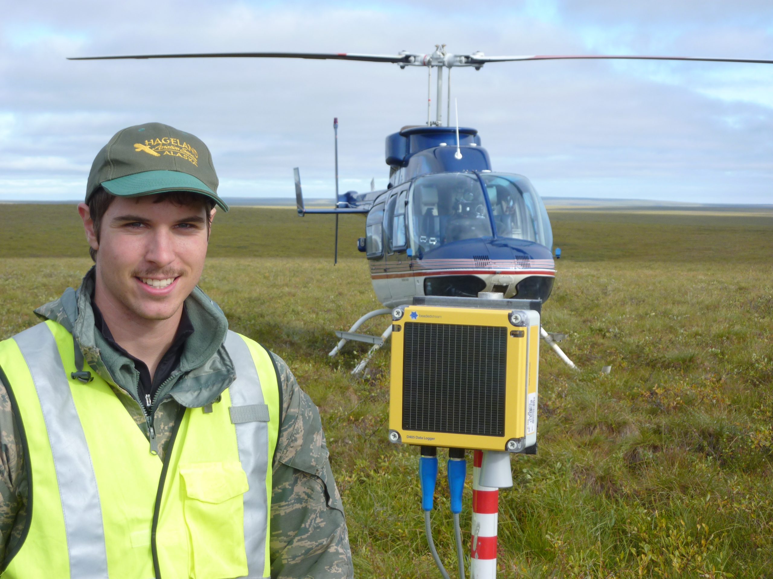 Beadedstream project manager holding a D405 datalogger in the Arctic tundra, helicopter in the background